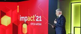 Wicepremier i minister rozwoju, pracy i technologii Jarosław Gowin podczas otwarcia wiosennej edycji Impact"21, po jego lewej stronie logo Impact'21 na czerwonym tle