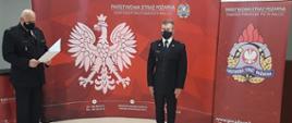 Zdjęcie przedstawia funkcjonariuszy PSP na czerwony tle z białym orłem w koronie i logo PSP w trakcie odczytu aktu powierzenia obowiązków zastępcy dowódcy zmiany