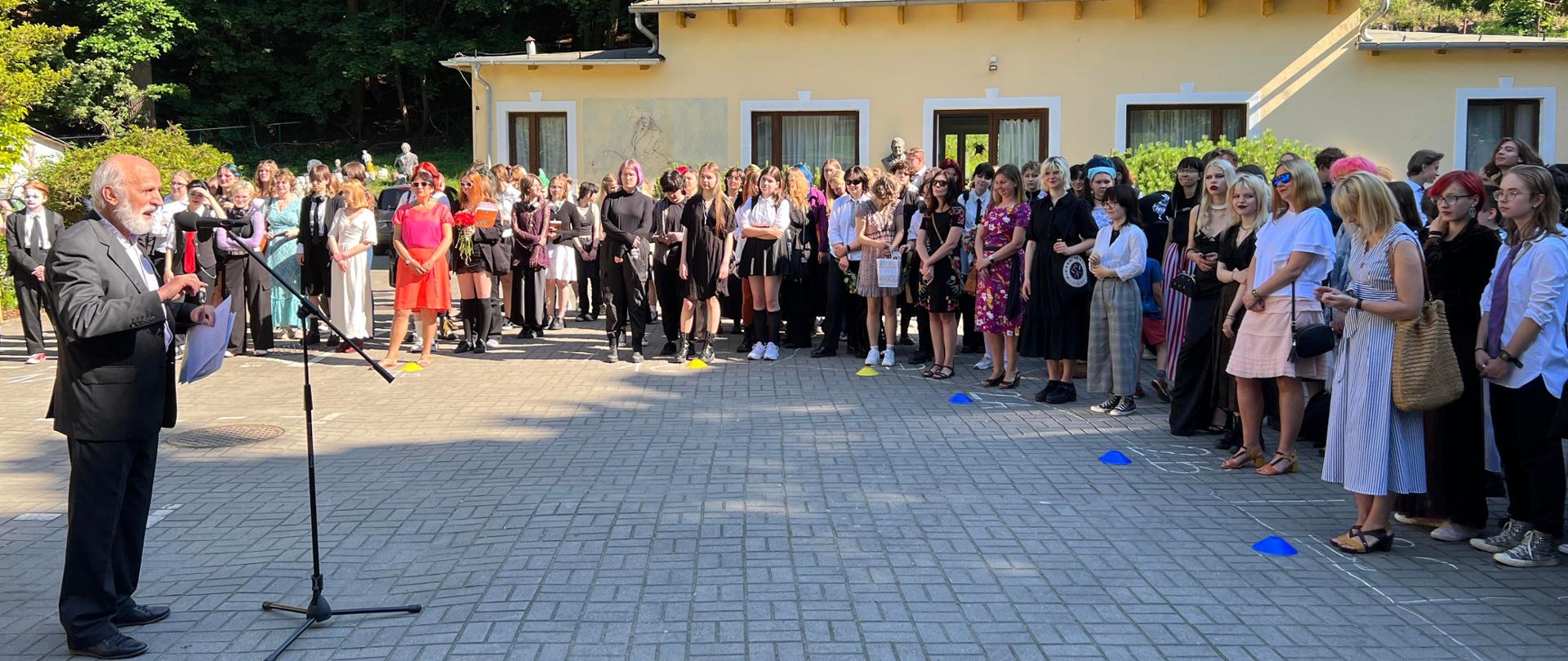 Zakończenie roku szkolnego w ZSP w Gdyni. Plac przed budynkiem szkoły. Dyrektor przemawia przez mikrofon do dużej grupy uczniów.