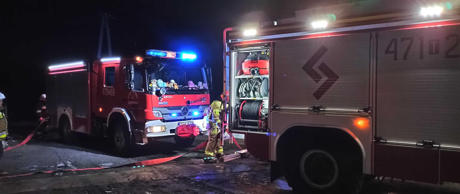 Opis Alternatywny: zdjęcie zrobione w nocy. Na zdjęciu widać dwa samochody strażackie i rozwinięte węże strażackie. Obok samochodów strażackich widać dwóch strażaków, którzy obsługują samochody.