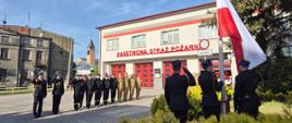 Na pierwszym planie znajduje się grupa strażaków w galowych, ciemnych mundurach, salutujących polskiej fladze powiewającej na wietrze. Strażacy stoją w rzędzie na placu przed budynkiem straży pożarnej. Budynek jest jasny, z czerwonymi bramami garażowymi i napisem "PAŃSTWOWA STRAŻ POŻARNA" umieszczonym nad nimi. Na budynku widoczne są także czerwone pasy. Za strażakami i budynkiem widać fragmenty innych budynków, w tym jeden z wieżą, oraz błękitne niebo z delikatnymi chmurami. Po prawej stronie kadru rosną zielone krzewy. Całość sceny sugeruje uroczystość lub apel strażacki.