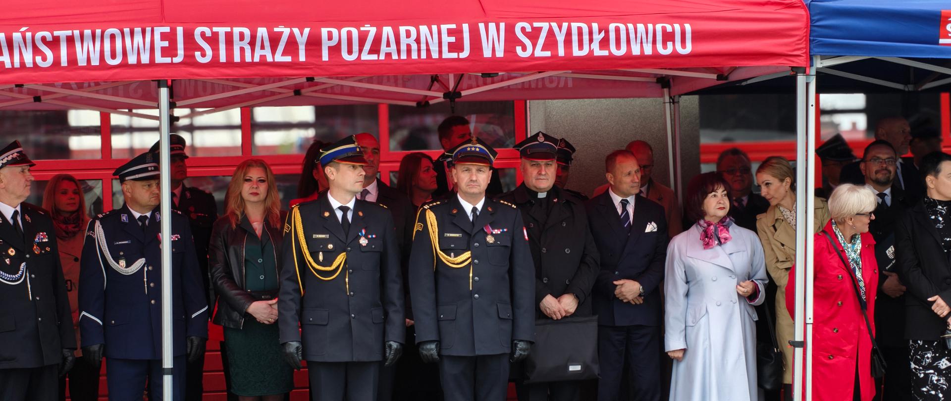 Na pierwszym planie dwóch strażaków PSP w czarnych mundurach. Wokół nich inni uczestnicy uroczystości. Stoją pod czerwonym namiotem. 