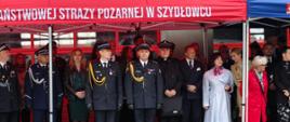 Na pierwszym planie dwóch strażaków PSP w czarnych mundurach. Wokół nich inni uczestnicy uroczystości. Stoją pod czerwonym namiotem. 