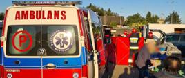 Po lewej stronie widoczny tył ambulansu z literą P na tylnych drzwiach. Po prawej stronie przed ambulansem stoi strażak trzymając parawan koloru czerwonego. Za parawanem widoczna głowa strażaka w hełmie. Po prawej stronie osoby postronne na targu z zamazanymi twarzami.