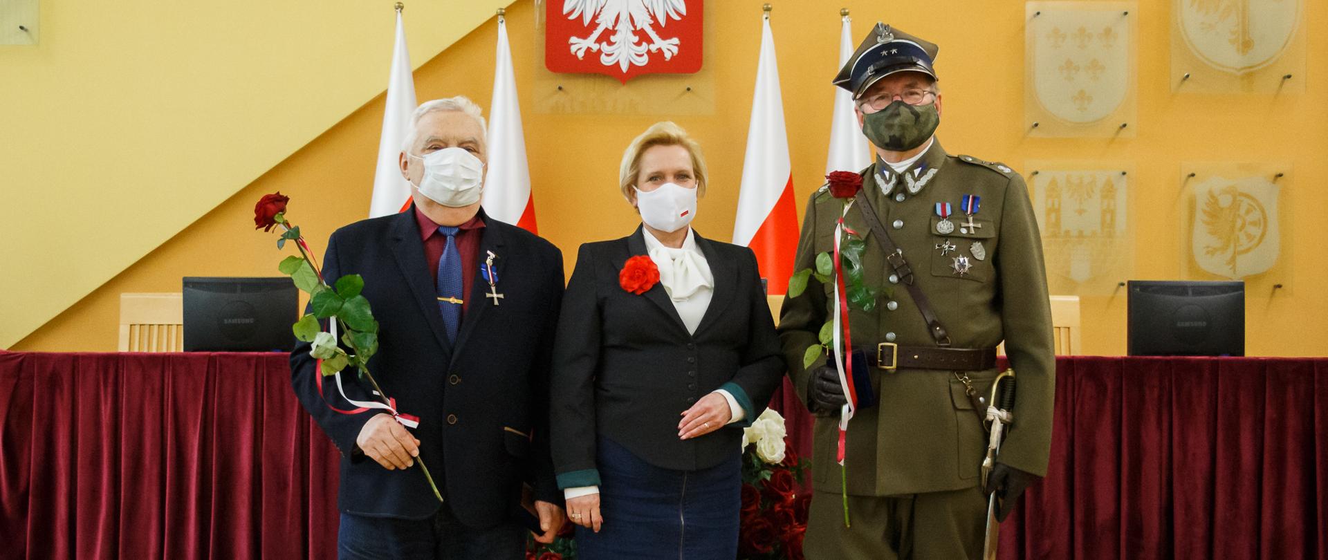 Na zdjęciu trzy osoby, ustawione centralnie do obiektywu - pozują do zdjęcia. Od lewej starszy mężczyzna w siwych włosach, trzymający czerwoną różę. W środku wicewojewoda opolski ubrana w granatową spódnicę ,czarny żakiet i białą koszulę. Na jednej z klap żakietu widać broszkę w postaci czerwonego kwiatka. Obok wicewojewody stoi wysoki mężczyzna ubrany w strój ułana. On również trzyma w dłoni czerwoną różę, w drugiej ręce trzyma szablę. Za trójką osób widać stół prezydialny, przykryty masywną, ciemną tkaniną. W dalszym planie widać sześć ustawionych równo obok siebie flag RP oraz wiszące na ścianie godło. 
