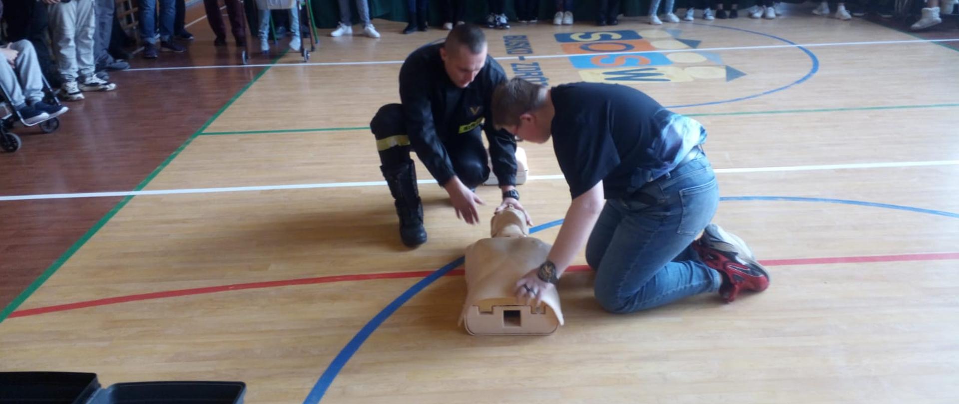 Strażak PSP oraz uczeń przy manekinie. Uczeń ćwiczy udzielanie pierwszej pomocy. W tle ludzie, a nad nimi zwisające kotary