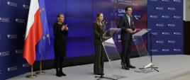 Minister Katarzyna Pełczyńska-Nałęcz and Deputy Minister Jan Szyszko stand on stage in front of microphones. To the left is a sign language interpreter and the Polish and EU flags