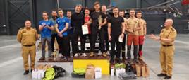 II Ogólnopolskie Zawody Grup Ratownictwa Wysokościowego - zdobywcy trzech pierwszych miejsc na podium - reprezentanci Podlasia - czterech strażaków z grupy ratownictwa wysokościowego na trzecim miejscu. Zdjęcie zrobione na hali, na której odbyły się zawody. Obok podium stoją torby z upominkami, a także dwóch Komendantów w mundurach koloru żółtego. Jeden z nich bije brawo.