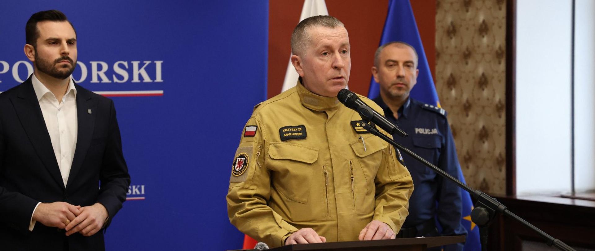 Na zdjęciu widać konferencję prasową w pomieszczeniu urzędowym. W centrum stoi mężczyzna w jasnobrązowym mundurze służbowym, przemawiający do mikrofonu przy mównicy z godłem Polski. Ma krótkie włosy i poważny wyraz twarzy. Za nim stoją dwaj inni mężczyźni: po lewej w ciemnym garniturze i białej koszuli, po prawej w granatowym mundurze policyjnym. W tle znajdują się flagi Polski i Unii Europejskiej oraz niebieska tablica z napisem urzędowym. Cała scena ma oficjalny, formalny charakter.