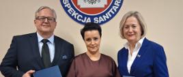 Osoby stojące na tle jasnej ściany. Nad nimi wisi logo Państwowej Inspekcji Sanitarnej