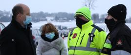 Grupa czterech osób: pierwszy z lewej Wojewoda obrany w ciemną kurtkę, w jasnej maseczce, bez nakrycia głowy. Druga osoba - przedstawicielka Wojewody, ubrana w jasną kurtkę, w jasnej maseczce bez nakrycia głowy. Trzecia osoba - Wójt gminy ubrany w kurtkę i czapkę odblaskową, w czarnej maseczce, ostatni - komendant powiatowy PSP ubrany w kurtkę strażacką, ciemną czapkę i maseczkę. 