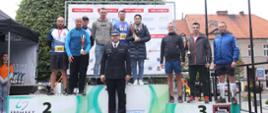 Zdjęcie grupowe - na podium w strojach sportowych stoi grupa 8 mężczyzn i kobieta trzymająca puchar, przed podium stoi mężczyzna w mundurze wyjściowym PSP