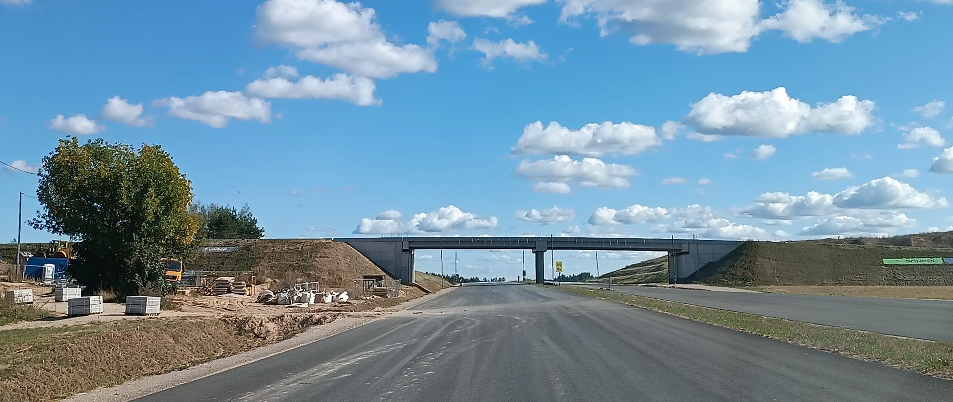 Widok na nowo wybudowaną drogę asfaltową prowadzącą do wiaduktu nad trasą, pod niebem z rozproszonymi białymi chmurami. Po lewej stronie widoczne są materiały budowlane i drzewo, po prawej nasypy ziemne.