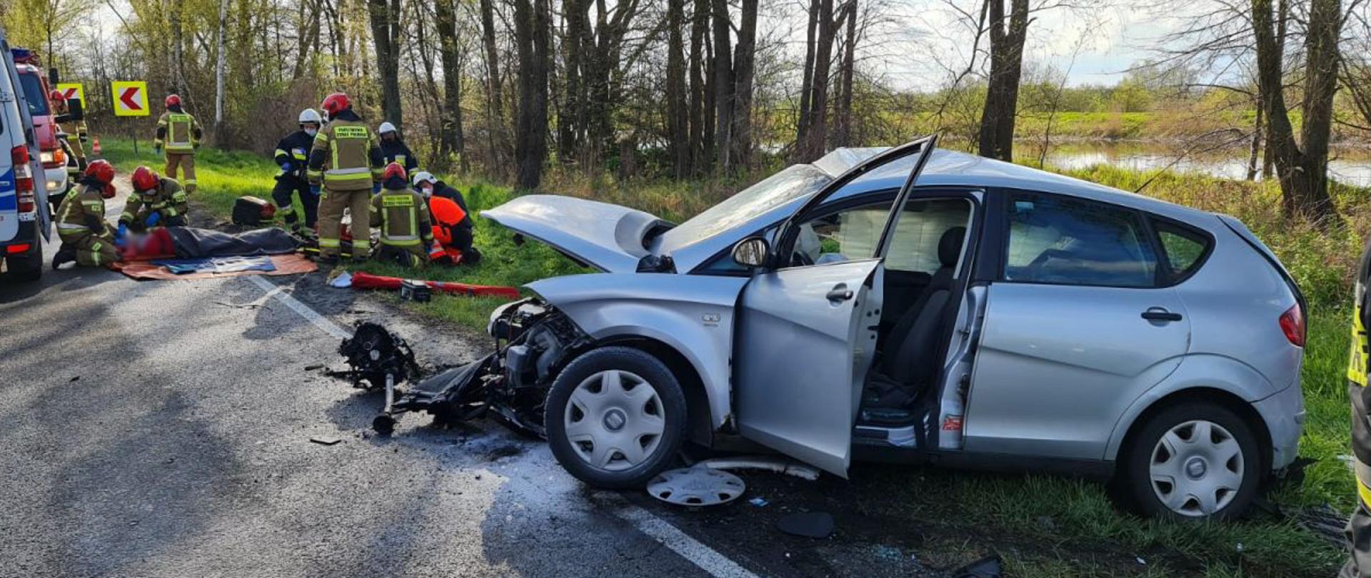 Na pierwszym planie znajduje się rozbity pojazd marki Seat z widocznie uszkodzonym przodem. Po lewej stronie kilku strażaków udziela kwalifikowanej pierwszej pomocy osobie poszkodowanej w wypadku.