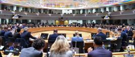 Wiceminister Kamila Król na Radzie UE ds. konkurencyjności. Na zdjęciu widać salę, na której obradują uczestnicy spotkania. Uczestnicy siedzą przy dużym okrągłym stole.