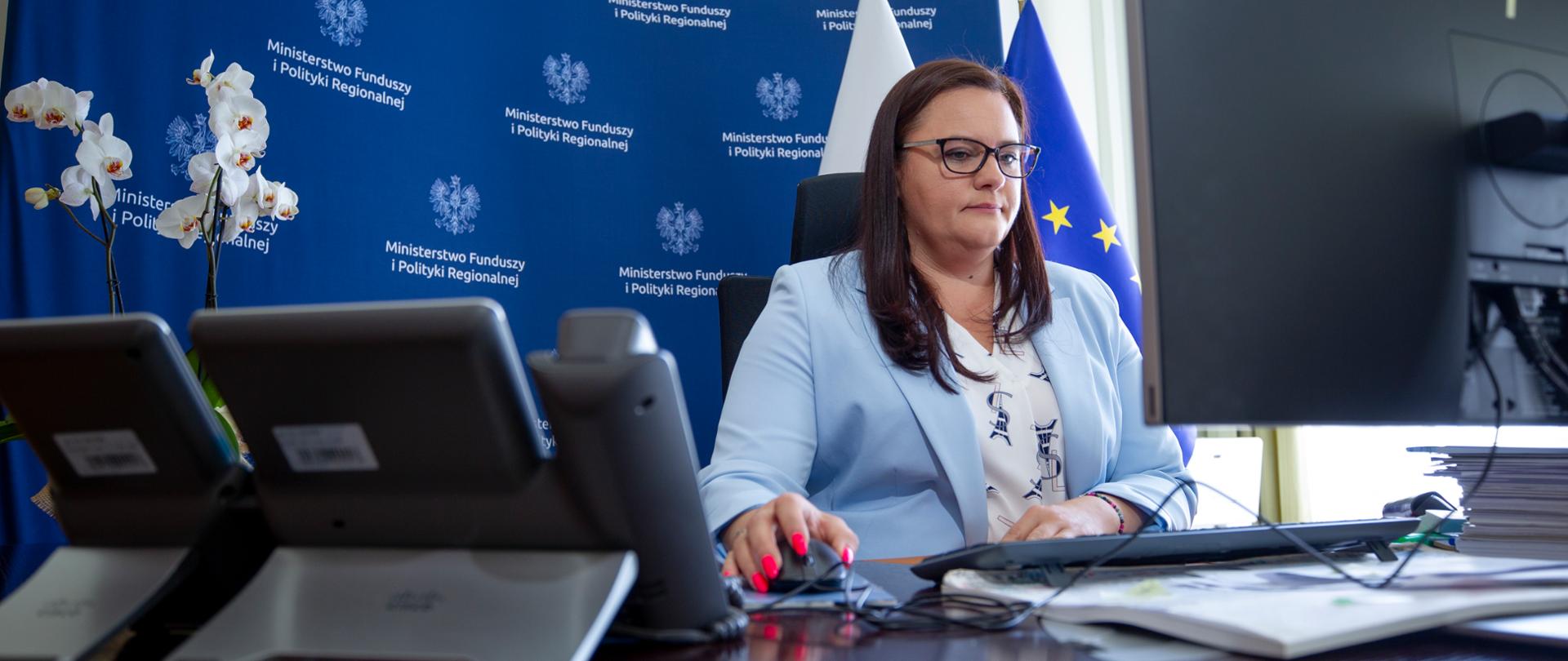 W gabinecie przy biurku z monitorem siedzi wiceminister Małgorzata Jarosińska-Jedynak. Za nią ścianka konferencyjna z logo Ministerstwa Funduszy i Polityki Regionalnej.