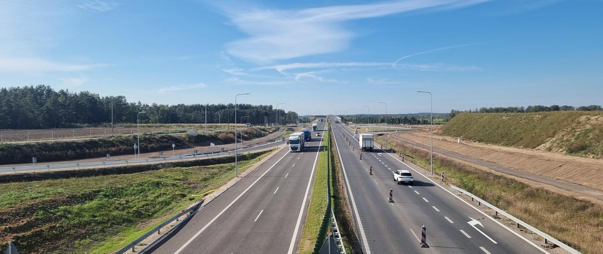 Na zdjęciu widać jezdnię, którą oddziela pas zieleni. Po jezdni jadą samochody osobowe i ciężarowe. Po lewej stronie widać las.