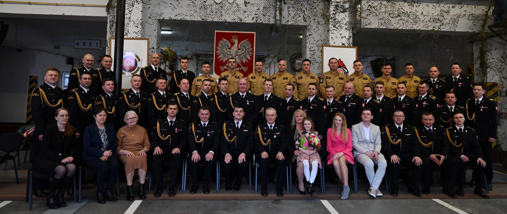 Kolorowa fotografia wykonana w garażu strażnicy PSP w Brzozowie. Przedstawia strażaków i pracowników cywilnych ubranych w mundurach wyjściowych i służbowych na wspólnym zdjęciu z okazji przejścia na emeryturę zastępcy komendanta PSP w Brzozowie. W tle godło narodowe oraz logo PSP i Związku Ochotniczych Straży Pożarnych.
