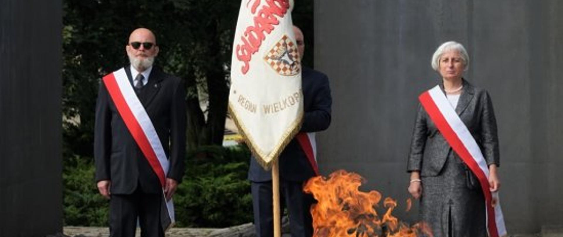 zdjęcie przestawia palący się znicz, za którym stoi poczet sztandarowy złożony z trzech osób. Po lewej stoi mężczyzna ubrany w szary garnitur, białą koszulę i krawat, przez ramię ma przewieszoną szarfę biało-czerwoną, w środku stoi mężczyzna trzymający sztandar. Obok stoi kobieta w szarej garsonce z przewieszoną przez ramię biało-czerwoną szarfą. 