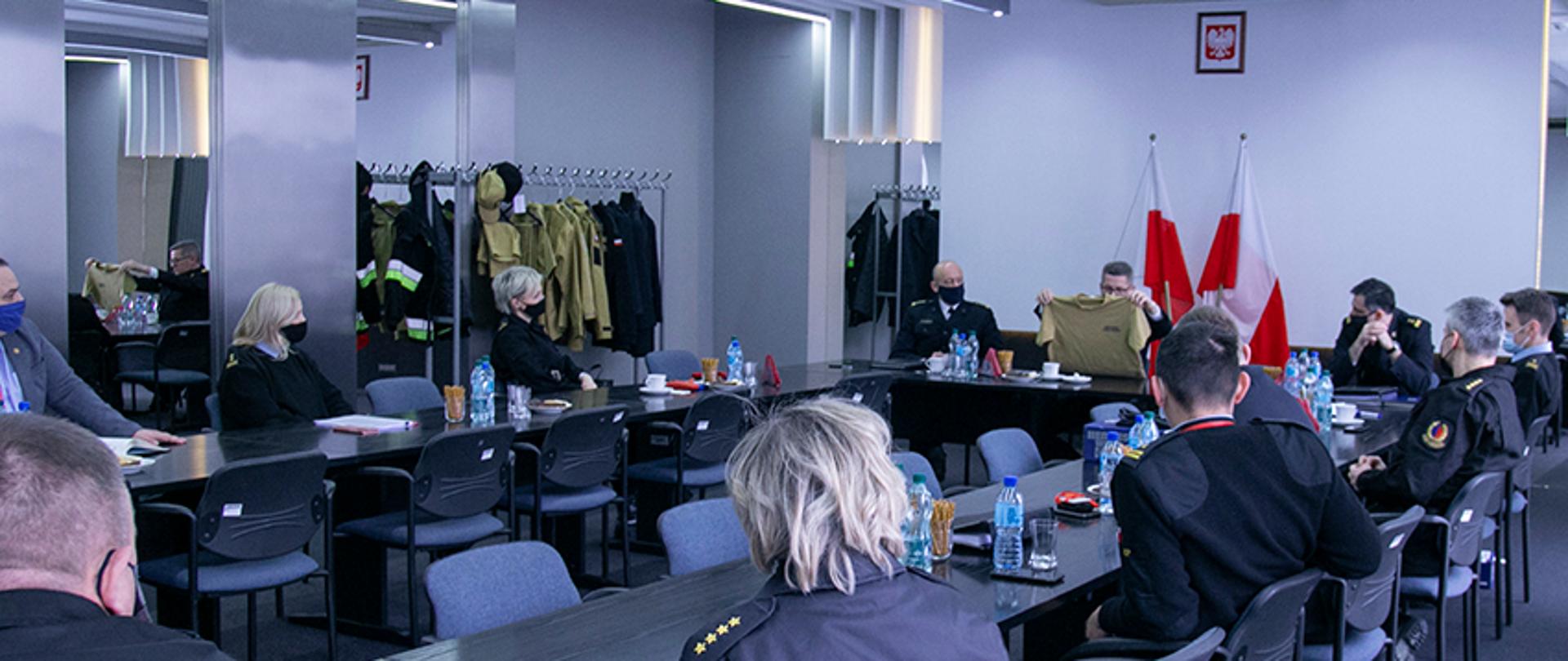 Zespół mundurowy podczas obrad. Wszyscy uczestnicy spotkania w maseczkach ochronnych. Przewodniczący prezentuje jeden z elementów nowego unundurowania