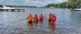 Na zdjęciu widać pięciu ratowników wodnych – członków ochotniczych straży pożarnych stojących po kolana w wodzie jeziora Trzesiecko. Ubrani są w specjalistyczne stroje ratownicze – kombinezony w kolorach czerwonym, pomarańczowym i zielono-czarnym oraz czerwone kaski ochronne. Wszyscy mają na sobie kamizelki asekuracyjne. W tle widoczne są jachty i łodzie zacumowane przy pomostach oraz zalesione brzegi jeziora. Pogoda jest umiarkowana – niebo jest częściowo zachmurzone, ale widać również przebijające się słońce. Zdjęcie zrobione podczas szkolenia służb ratowniczych na wodzie.