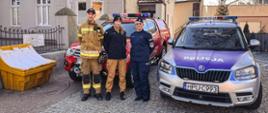 Dwóch strażaków i policjant stoją przy samochodach służbowych przed budynkiem krwiodawstwa i krwiolecznictwa.