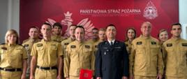 Grupa kilkunastu funkcjonariuszy i pracowników Państwowej Straży Pożarnej pozująca do wspólnego zdjęcia na tle czerwonej ścianki reklamowej komendy powiatowej psp w wołowie. Zdecydowana większość osób ubrana jest w piaskowe (beżowe) mundury służbowe i specjalne. W centrum stoi mężczyzna w ciemnogranatowym mundurze wyjściowym, a obok niego strażak w piaskowym mundurze trzymający czerwoną teczkę z logo straży. W tle widoczna jest czerwona ścianka z dużym, białym godłem Polski, logo psp oraz napisem państwowa straż pożarna komenda powiatowa psp w wołowie.