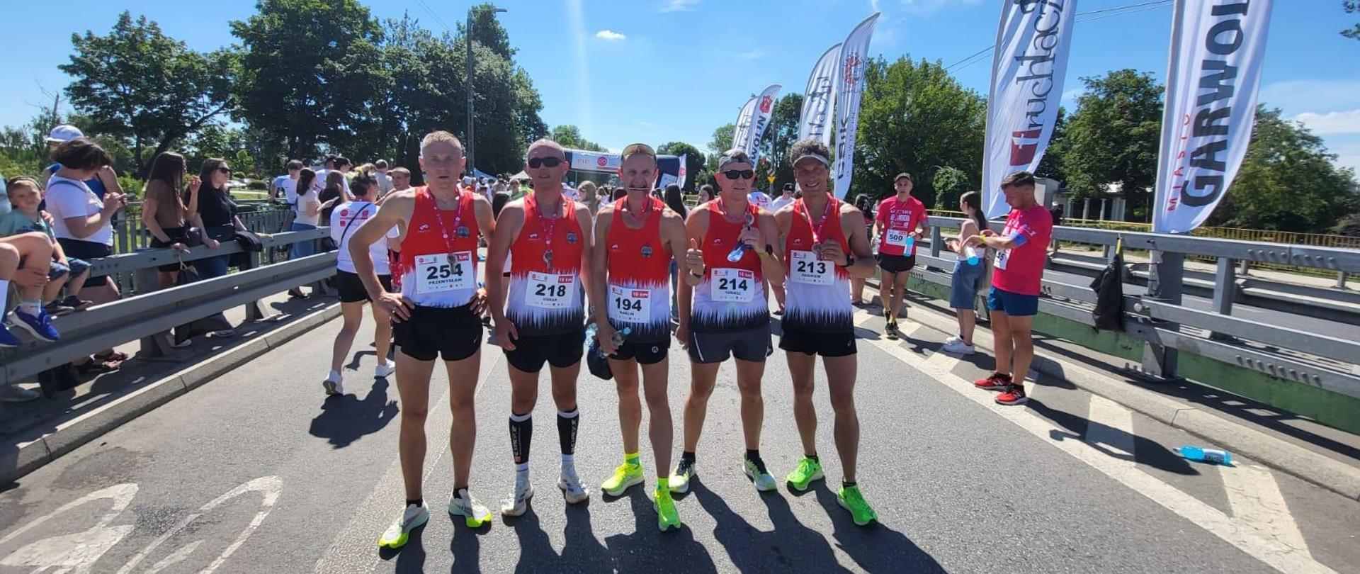 Pięciu zawodników w strojach biegaczy pozujących do zdjęcia na ulicy. W tle kibice zawodów.