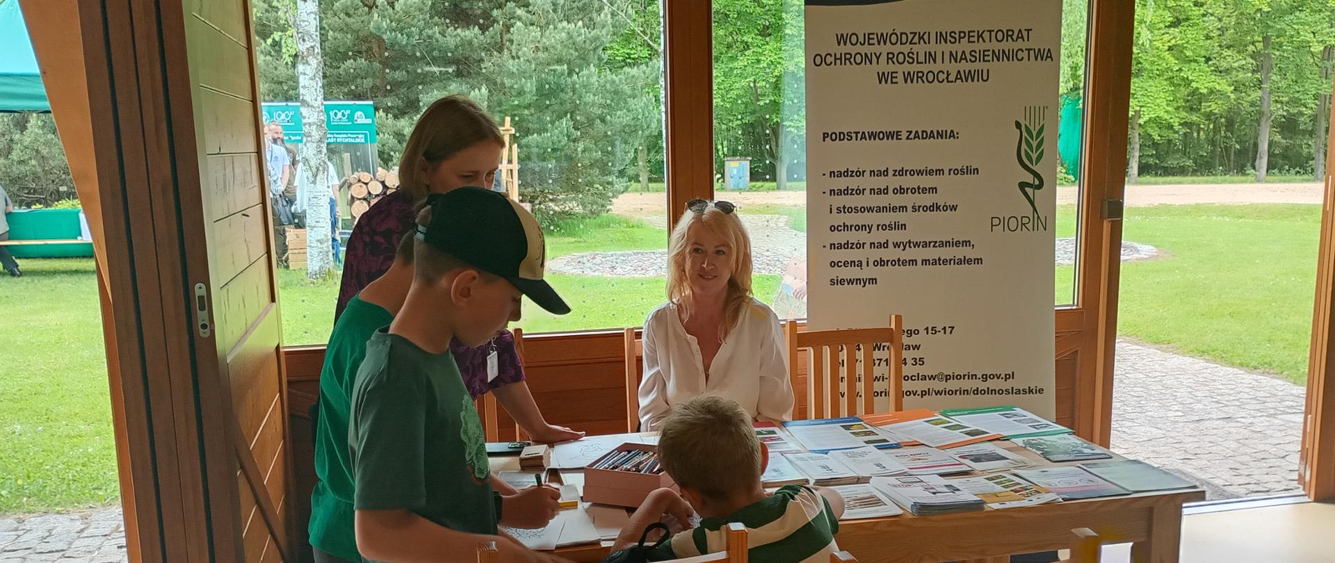 Przy drewnianym stole w przeszklonym pawilonie siedzą i stoją dzieci oraz dwie panie inspektor. Na stole znajdują się materiały informacyjne i karty do wypełnienia. W tle widoczny jest baner Wojewódzkiego Inspektoratu Ochrony Roślin i Nasiennictwa we Wrocławiu oraz zielony teren za oknami.