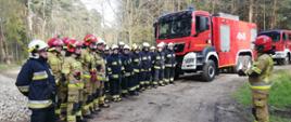 Grupa strażaków PSP i OSP ustawiona w szeregu uczestniczy w odprawie prowadzonej przez dowódcę ćwiczeń. Strażacy mają na sobie ubrania bojowe. W tle pojazdy pożarnicze uczestniczące w ćwiczeniach. 