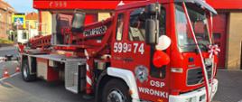 Plac przed remizą OSP Wronki. Na tle budynku remizy stoi samochód specjalny z podnośnikiem hydraulicznym. Przystrojony jest w balony i biało-czerwone wstęgi. Na drzwiach widnieje napis OSP Wronki oraz herb Wronek.