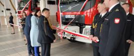 Przed samochodem pożarniczym strażak ubrany w mundur wyjściowy oraz dwie osoby w ubraniu cywilnym przecinają wstęgę koloru biało-czerwonego.