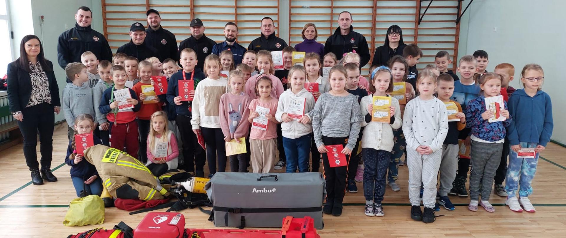 Zdjęcie zrobione na sali gimnastycznej. Na zdjęciu ustawione obok siebie kilkanaście dzieci, za nimi nauczycieli i kilku strażaków. Na dole zdjęcia leżą czerwone torby medyczne strażaków.