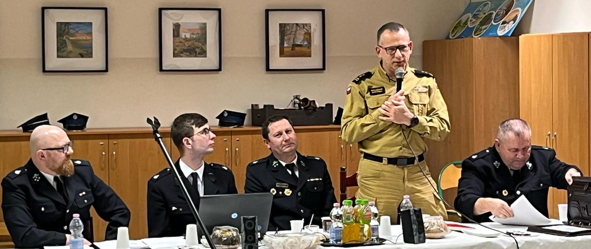Grupa mężczyzn siedzi w oświetlonej sali za stołem prezydialnym, jeden mężczyzna ubrany w mundur straży pożarnej przemawia na stojąco z mikrofonem w ręku