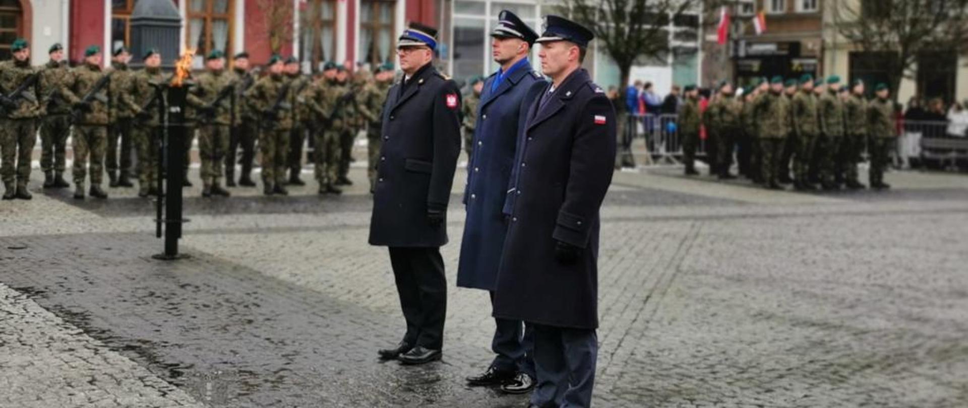 Zbiórka żołnierzy stojących w dwuszeregu na utwardzonym placu. Na pierwszym planie stoi trzech funkcjonariuszy w ciemnych płaszczach. W tle kolorowe fasady kamienic.