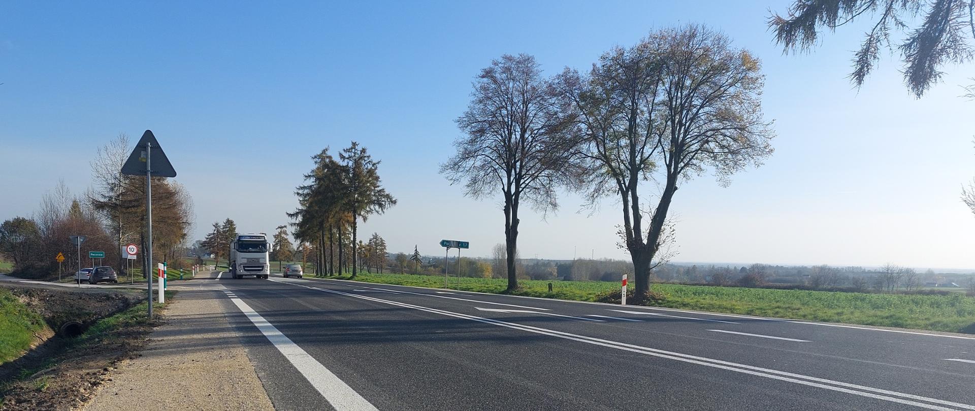 DK73 Pacanów - jednojezdniowa droga asfaltowa, w oddali samochód ciężarowy i osobowy, z prawej strony wzdłuż drogi pobocze z kruszywa, rów, przepust, skrzyżowanie, drzewa przy drodze 