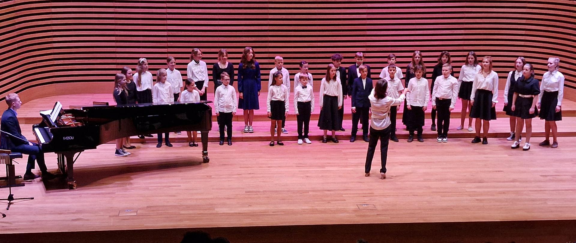 Dzieci stoją w dwóch rzędach i śpiewają, z przodu na środku dyryguje kobieta, po lewej stronie siedzi mężczyzna przy fortepianie.