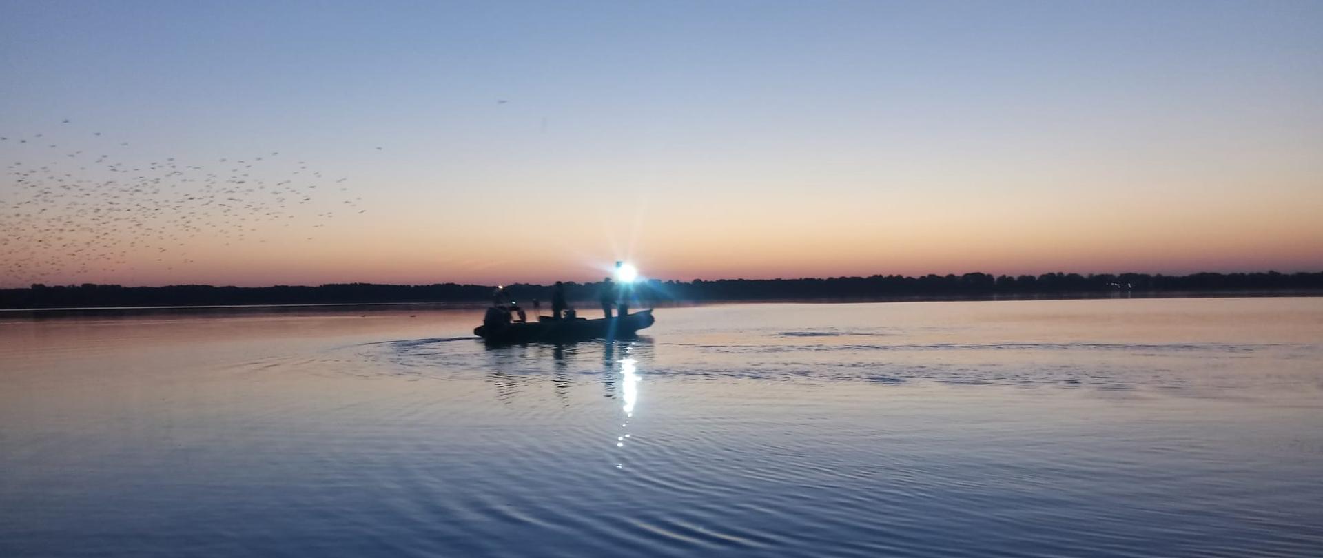 Strażacy płyną łódką strażacką. Pora wieczorna. Na horyzoncie widać drzewa.