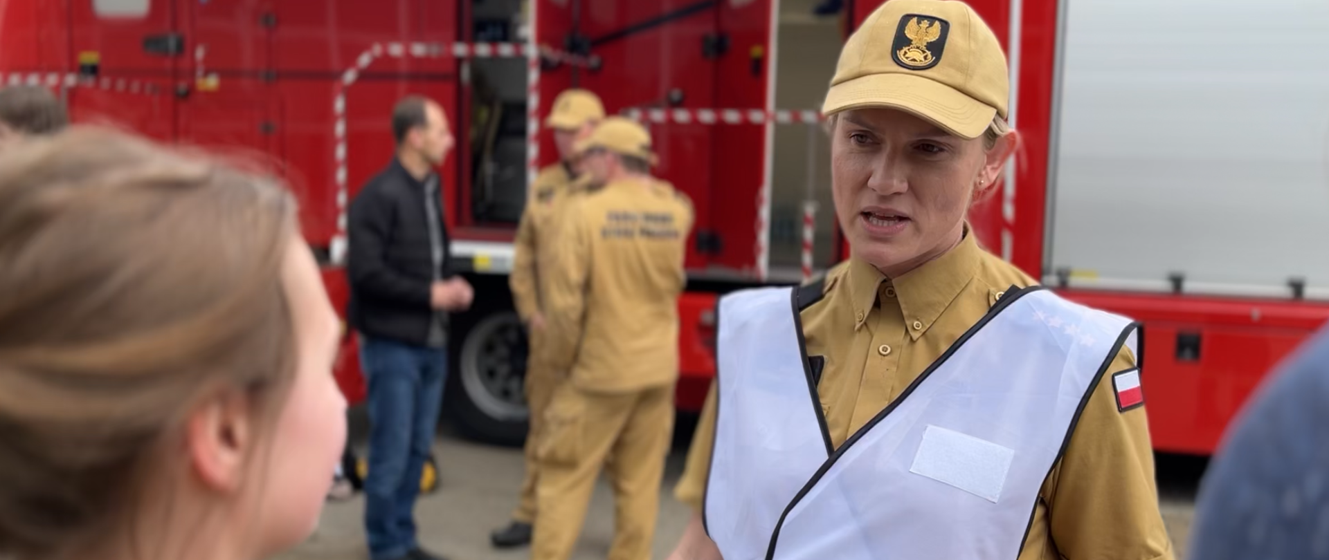 Kobieta strażak, dodatkowo w białej kamizelce. Z lewej strony widać dziewczynę, rozmawiają. W dali grupka strażaków, w tle samochód pożarniczy.