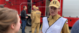 Kobieta strażak, dodatkowo w białej kamizelce. Z lewej strony widać dziewczynę, rozmawiają. W dali grupka strażaków, w tle samochód pożarniczy.