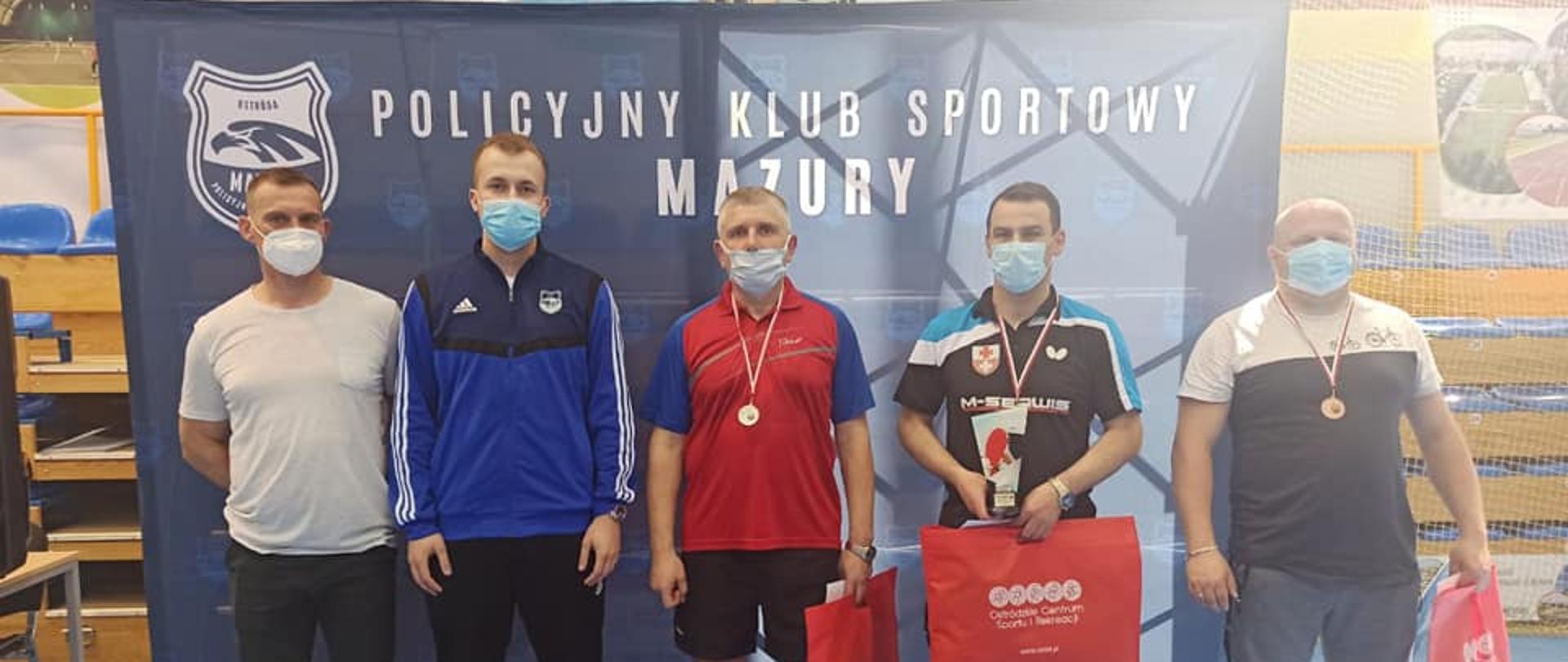 Na zdjęciu stoi obok siebie pięciu zawodników ubranych w stroje sportowe. Troje z nich ma szyi medale. Za nimi znajduje się duży baner z napisem Policyjny Klub Sportowy Mazury.
