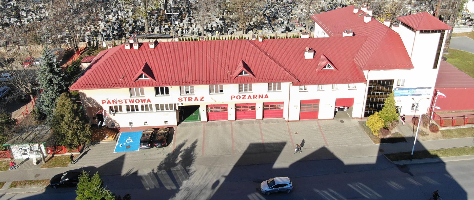 Na fotografii przedstawiono budynek komendy. Zdjęcie zrobione z lotu ptaka. Widoczne niebieskie miejksca parkingowe tuż przed samą komendą. Miesce oznakowane białą ikoną człowieka na wózku inwalidzkim.