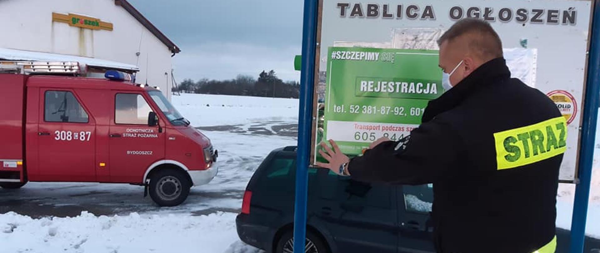 Na pierwszym planie strażak OSP stojący bokiem w czarnym ubraniu koszarowym. Strażak stoi przy tablicy ogłoszeń i przykleja do niej plakat informujący o szczepieniach. W tle samochód pożarniczy czerwony marki Lublin należący do OSP Bydgoszcz. 