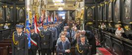 Zdjęcie przedstawia strażaków z Państwowej i Ochotniczej Straży Pożarnej w mundurach wyjściowych ze sztandarami, którzy znajdują się w Bazylice mniejszej p.w. św. Mikołaja Biskupa w Bochni . Po lewej stronie jeden za drugim stoją poczty sztandarowe na czele których znajduje się oficer z Państwowej Straży Pożarnej w mundurze galowej. Po lewej stronie zdjęcia w ławka siedzą uczestnicy uroczystości: strażacy, policjanci i osoby cywilne. W tle wystrój barokowy kościoła