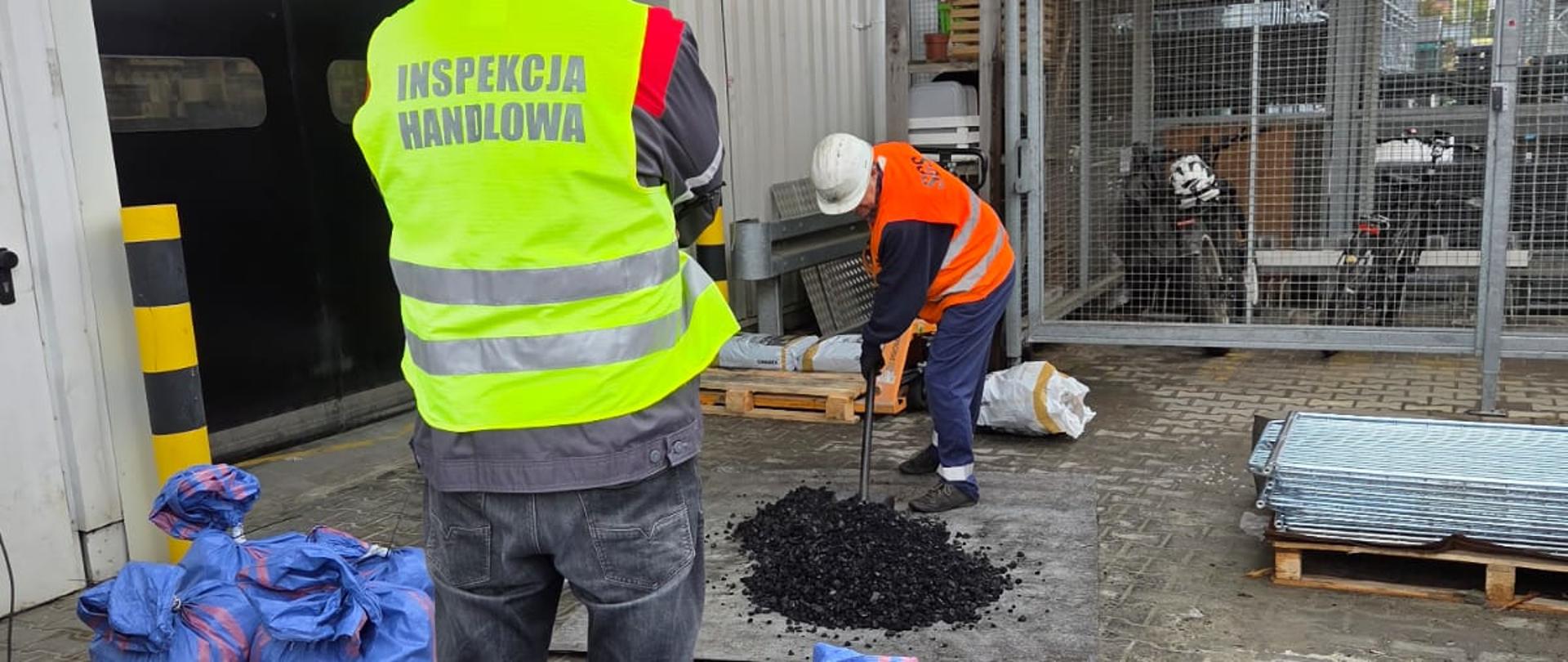 Na pierwszym planie niebieskie duże worki związane u góry, na środkowym planie stoi mężczyzna w kamizelce z napisem Inspekcja Handlowa, na dalszym planie mężczyzna z łopatą nabiera węgiel, który leży na plandece. 