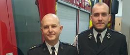 Dwóch strażaków w ciemnych mundurach galowych stoi w garażu pomiędzy czerwonymi wozami bojowymi. Mężczyzna po lewej trzyma białą planszę z czerwonym logo KSRG i napisem odnoszącym się do Komendy w Bytomiu.