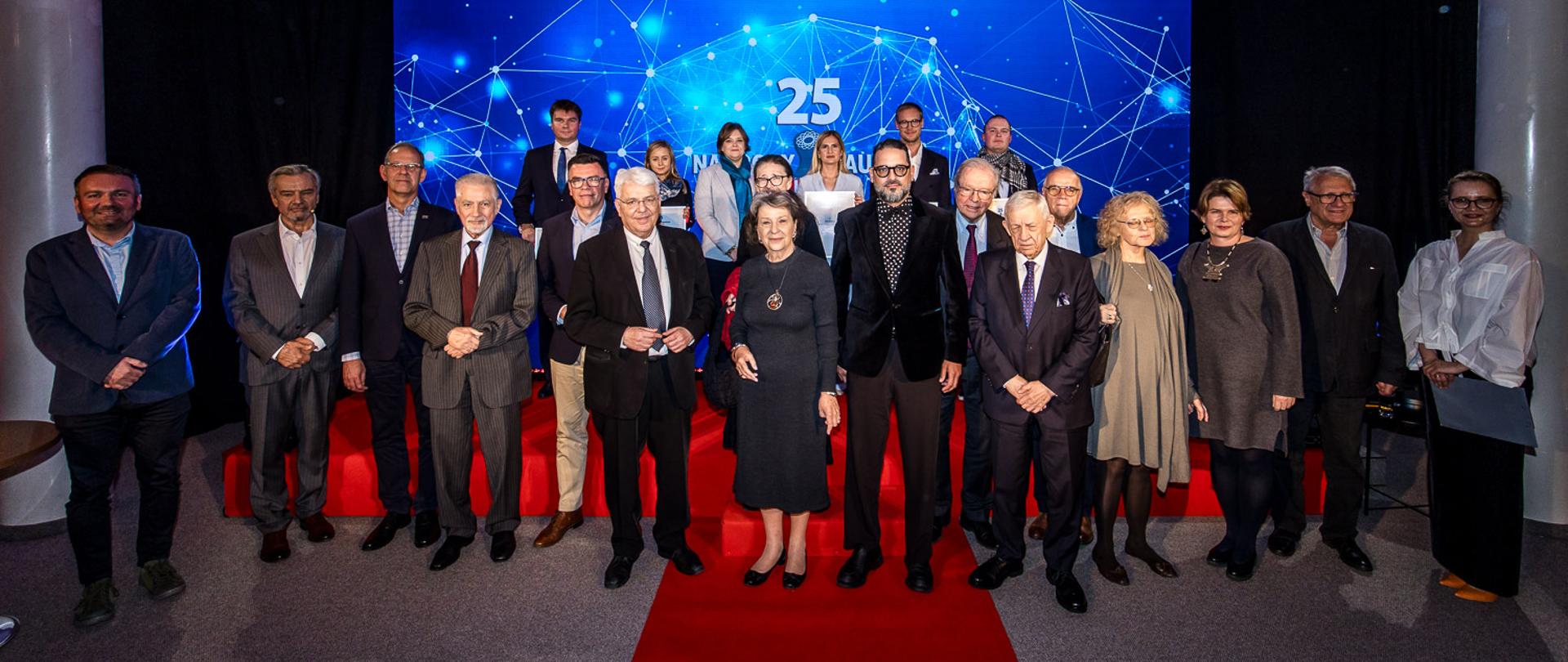 Zdjęcie zbiorowe, na szarej podłodze z czerwonym dywanem stoi grupa kilkudziesięciu osób, za nimi na ścianie ekran z napisem 25 na niebieskim tle.