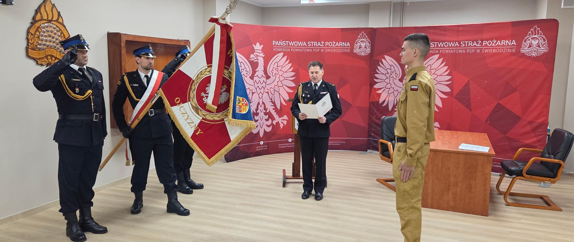Zdjęcie przedstawia akt składania ślubowania przez nowoprzyjętego funkcjonariusz na sztandar Komendy Powiatowej Państwowej Straży Pożarnej w Świebodzinie. Akt Ślubowania odczytuje Komendant Powiatowy brygadier Mariusz Wójcik.