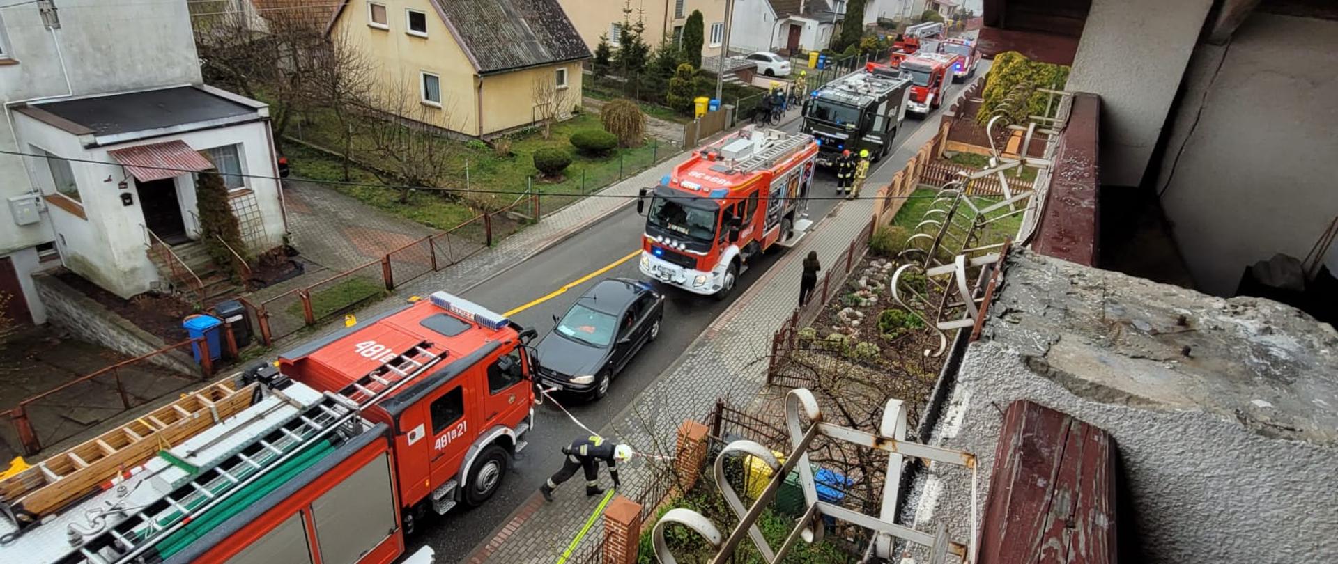 Fotografia przedstawia budynki mieszkalne oraz ulicę na, której znajdują się wozy strażackie i strażacy pracujący na miejscu zdarzenia.
