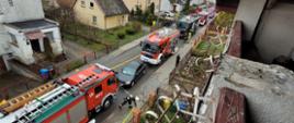 Fotografia przedstawia budynki mieszkalne oraz ulicę na, której znajdują się wozy strażackie i strażacy pracujący na miejscu zdarzenia.